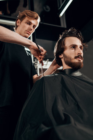 Barber works in barbershop. Handsome man with dark hair, beard and mustache has haircut in hairdressing saloon.の写真素材