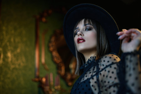 Attractive brunette girl in a black evening dress and black hat allures in a luxury vintage...