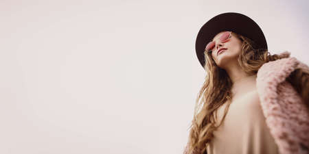 Autumn fashion. Stylish young woman in a fashionable fur coat and hat looks at the sky through pink glasses.の写真素材