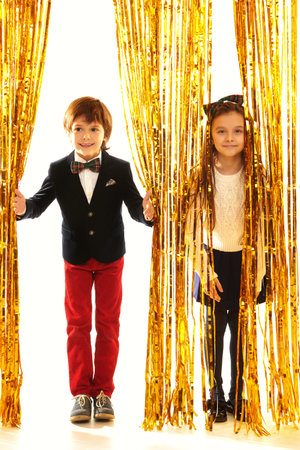 Children's party. Happy children in evening dresses peek out merrily from behind shiny gold foil curtains. Kid's fashion and holidays.の写真素材