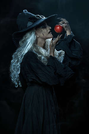Halloween. Portrait of an ugly old witch standing with her magic staff on a black background surrounded by sparks of fire. Witchcraft.の写真素材