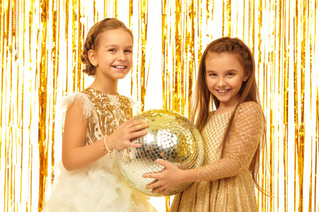 Kid's fashion and holidays. Beautiful child girl in a golden evening dress posing with disco ball on a background with shiny gold foil curtains. Children's party.の写真素材