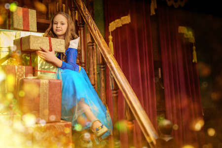 Christmas tale. A beautiful blonde girl in a princess dress sits on a wooden stairs with a lot of gifts and smiles dreamily. Vintage room.の写真素材