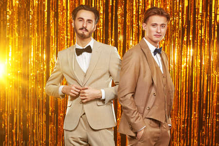 Christmas and New Year fashion. Two handsome young men in elegant evening suits stand on the background of shiny golden curtains. Men's fashion.の写真素材