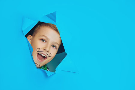Cool cheerful boy in a pirate costume peeks out of a hole in a blue background at the studio. Happy...
