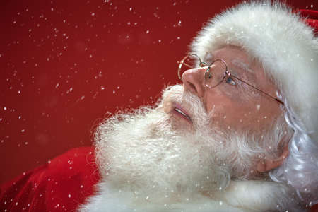 Close-up portrait of a jolly Santa Claus looking up over festive red background with snowfall. Merry Christmas and Happy New Year!の写真素材