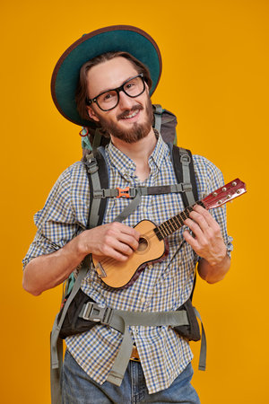 Funny smiling young man with a backpack and tourist hat holding a small guitar in his hands. Studio portrait in a yellow background. Tourism and travel, adventure.の写真素材