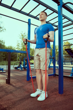 Handsome muscular man does strength exercises with elastic bands at outdoor gym. Active healthy...