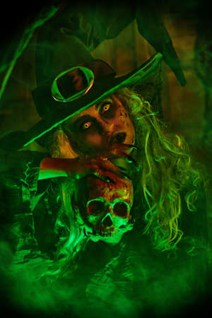 Halloween. A frightening old witch holds a skull in an old wooden house. Magical green and red light.の写真素材