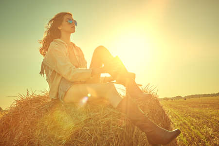 The spirit of freedom and adventure. A beautiful girl with wavy hair and in denim clothes sits in a field on a haystack against the backdrop of the setting sun. Wild west and hippie style. Copy space.の写真素材