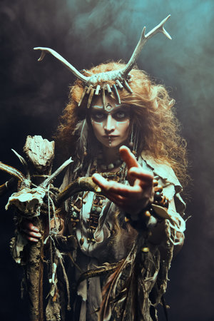 Female shaman in ethnic dress and deer antlers headdress surrounded by fog holds a ritual staff and...
