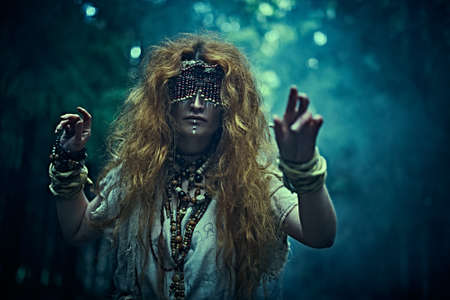 A frightening forest witch with a mask covering her eyes stands in a gloomy forest and holds out her arms forward. Woman shaman. Paganism. Death ritual. Halloween.の写真素材
