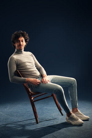 Full length portrait of a handsome young man with curly dark hair swinging on the chair on a black background. Men's beauty.の写真素材