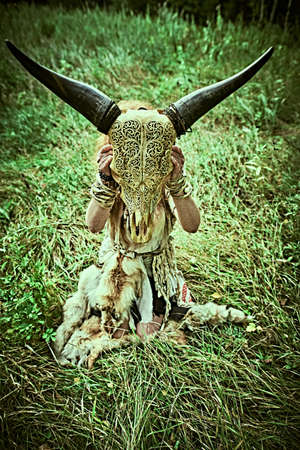 Female shaman in ethnic dress sits in a clearing in the forest and holds the skull of an animal in front of her. Mystical ritual. Fantasy concept, magic. Paganism. Halloween.の写真素材