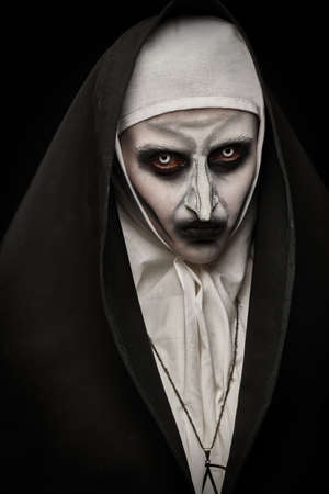 Halloween. Portrait of a scary devilish nun with bloody eyes looking at camera on a black background. Horrors.の写真素材