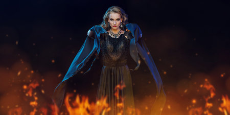 Portrait of a beautiful evil witch in an elegant dress conjuring among the flames of burning fire in...