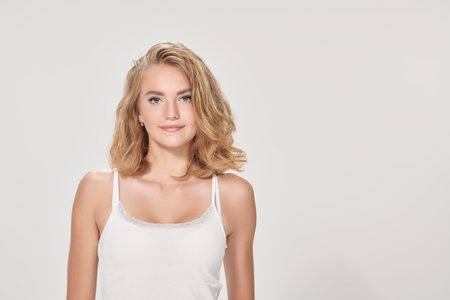 Studio portrait of a beautiful smiling blonde teenage girl in a white tank top posing in a white background with copy space. Youth style.の写真素材