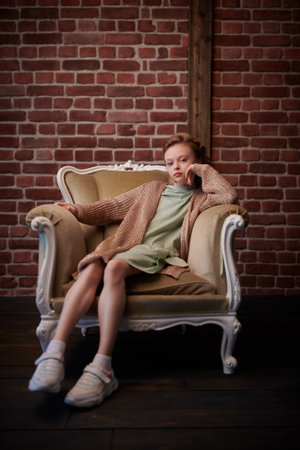 Kid's fashion. Full length portrait of a cute eight year old modern girl sitting in a vintage armchair in loft style room.の写真素材