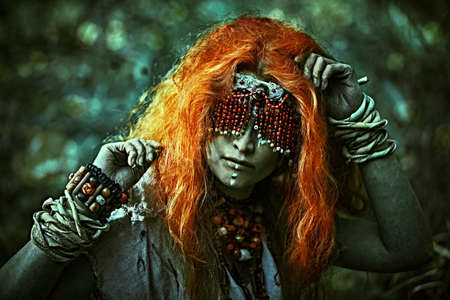 Frightening forest witch with a mask covering her eyes standing in a dark gloomy forest. Woman shaman. Paganism. Death ritual. Halloween.の写真素材