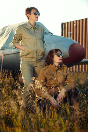 Full length portrait of two beautiful professional female commercial aviation pilots in stylish uniform and sunglasses with bright red lips posing on the airfield. Aviation.の写真素材