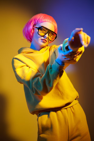 Modern girl with bright pink hair poses in yellow sunglasses and yellow hoodie suit in mixed color...