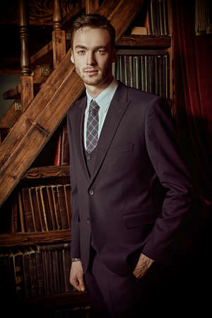 Men's beauty, fashion. Portrait of a handsome young man in a formal suit posing by the bookcase in a luxury apartment's interior.の写真素材
