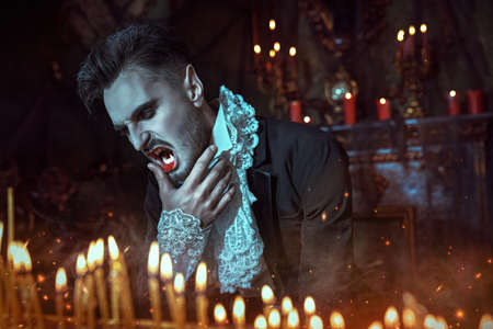 Bloodthirsty vampire baring his teeth among burning candles illuminating his dark gloomy castle at nighttime. Halloween.の写真素材