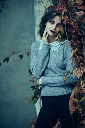 Halloween. Horror. A cute and terrible ghost of a girl with faded eyes stands at a ruined wall, shrouded in withered leaves, and smiles slightly.の写真素材