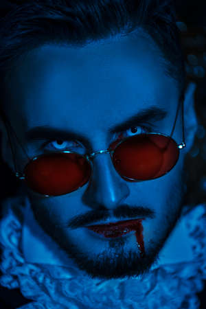 Close up portrait of a handsome blue-eyed vampire man with blood on his lips wearing an elegant 19th century suit and red sunglasses. Halloween. Vampires.の写真素材
