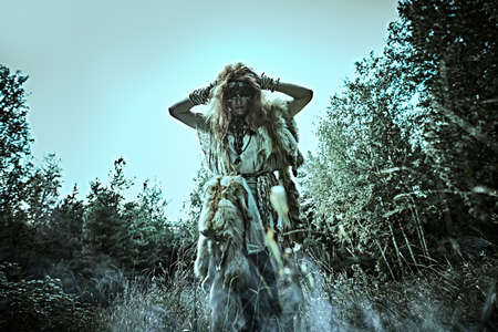 Mysterious shaman woman in a gloomy forest putting on a mask  for doing a magic ritual. Paganism. Death ritual. Halloween.の写真素材