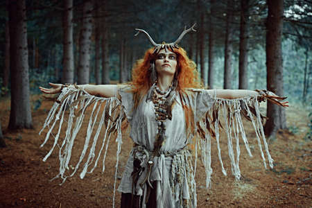 An ancient shaman woman in ritual garment performing a traditional shamanic ritual in the forest with her arms streched aside. Fairy forest witch. Fantasy. Halloween.の写真素材
