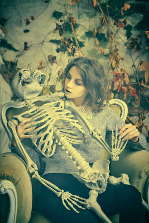 A pale ghost girl with dusty black hair and neat clothes sits in an old vintage chair with a skeleton in her hands against the background of a ruined vintage wall. Halloween. Horror.の写真素材