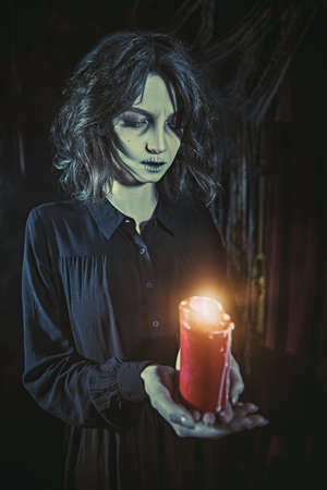 A lonely ghost girl in a black mourning dress stands with a candle in her hands in a dark gloomy room, shrouded in cobwebs. Horror film, thriller. Halloween.の写真素材