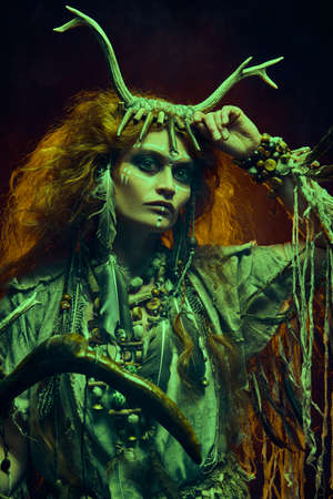 Portrait of a shaman woman in ritual clothes and deer antlers headdress posing in the dark background lit by fire behind her. Ethnic traditions. Paganism. Halloween.の写真素材