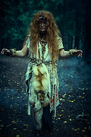 Woman shaman. Full length portrait of a frightening forest witch with a mask on her eyes doing her mystical ritual in a magic forest at night. Paganism. Death ritual. Halloween.の写真素材