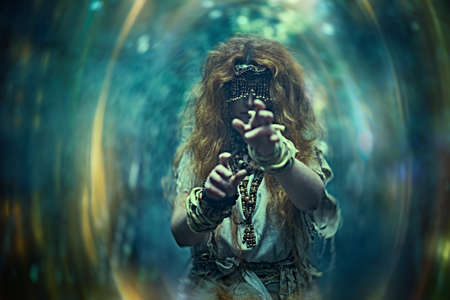 Scary forest witch with a mask covering her eyes in a dark gloomy forest creating a magic portal. Woman shaman. Paganism. Death ritual. Halloween.の写真素材