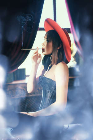 Sexual brunette woman in a black corset and stockings wearing a red hat smokes a cigarette in an expensive vintage apartment. Luxury lifestyle.の写真素材