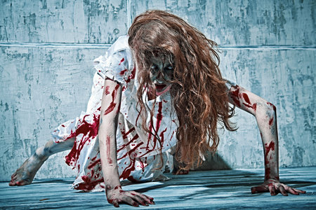 A creepy, devil-possessed woman with pale eyes and tousled hair covering her face and a bloody nightie crawls against a concrete wall. Zombie woman. Horror, thriller. Halloween.の写真素材