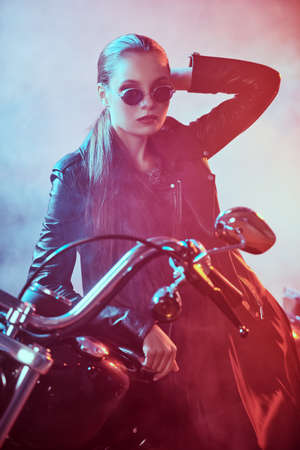 Confident girl biker in black leather jacket and black sunglasses stands in a garage leaning against her motorcycle.の写真素材