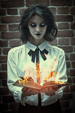 Portrait of a poltergeist girl with colorless eyes standing in a gloomy room by a brick wall with an opened burning book. Halloween. Horror scene.の写真素材