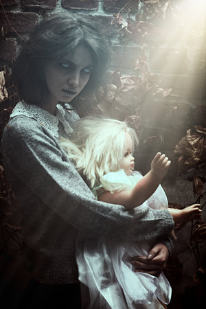 A frightening ghost woman with faded eyes stands with a baby doll among withered foliage by a ruined wall with strange light from above. Horror film, thriller. Halloween.の写真素材