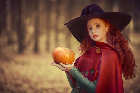 Halloween fairy tales. Beautiful young girl witch poses in the autumn forest with a pumpkin in her hands. Copy space.の写真素材