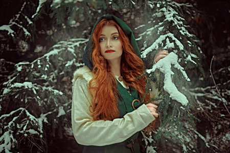Christmas tales. Pretty young woman with long red hair wearing medieval clothes walks in a snowy winter forest. Historical reconstruction of the early Middle Ages.の写真素材