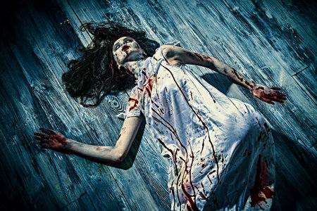 A scary bloody woman, possessed by the devil, bends in bloody nightgown on a floor in a dark room. Creepy zombie woman. Horror, thriller. Halloween.の写真素材