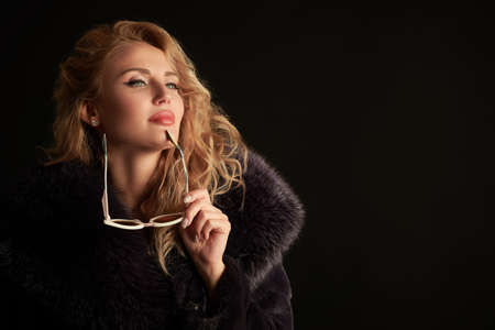Gorgeous blonde woman with curly blonde hair and elegant makeup poses in a luxurious black fur coat. Black studio background with copy space.  Fur coat fashion.の写真素材