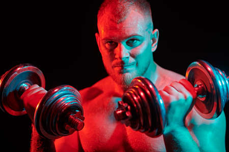 Portrait of a strong healthy handsome athletic man training hard with dumbbells on a black background with red light. Male beauty concept. Fitness, bodybuilding.の写真素材