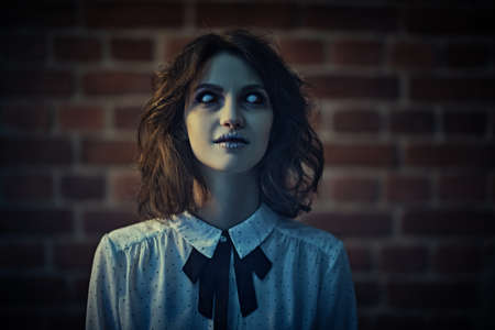 Horror scene. Portrait of a spooky ghost girl with faded eyes in a gloomy brick wall room. Halloween.の写真素材