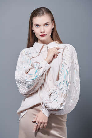 Beautiful girl fashion model with elegant makeup posing in a romantic beige blouse with wide sleeves and a beige straight skirt. High-fashion shoot. Studio portrait on a grey background.の写真素材