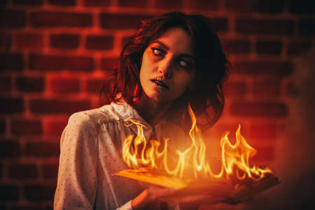 Vicious ghost girl with faded eyes stands with a burning book in her hands in a gloomy room lit by sinister red light. Horror scene. Halloween.の写真素材