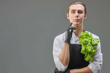 Portrait of a sophisticated male chef cook in uniform standing with lettuce leaves folded behind an apron and smiling slightly. Studio portrait on a grey background with copy space.の写真素材